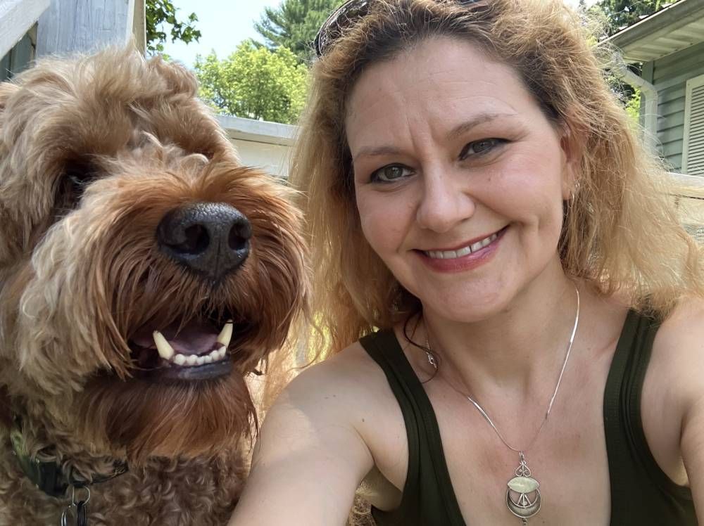 A smiling person poses for a selfie outdoors next to a shaggy, brown dog with its mouth open.