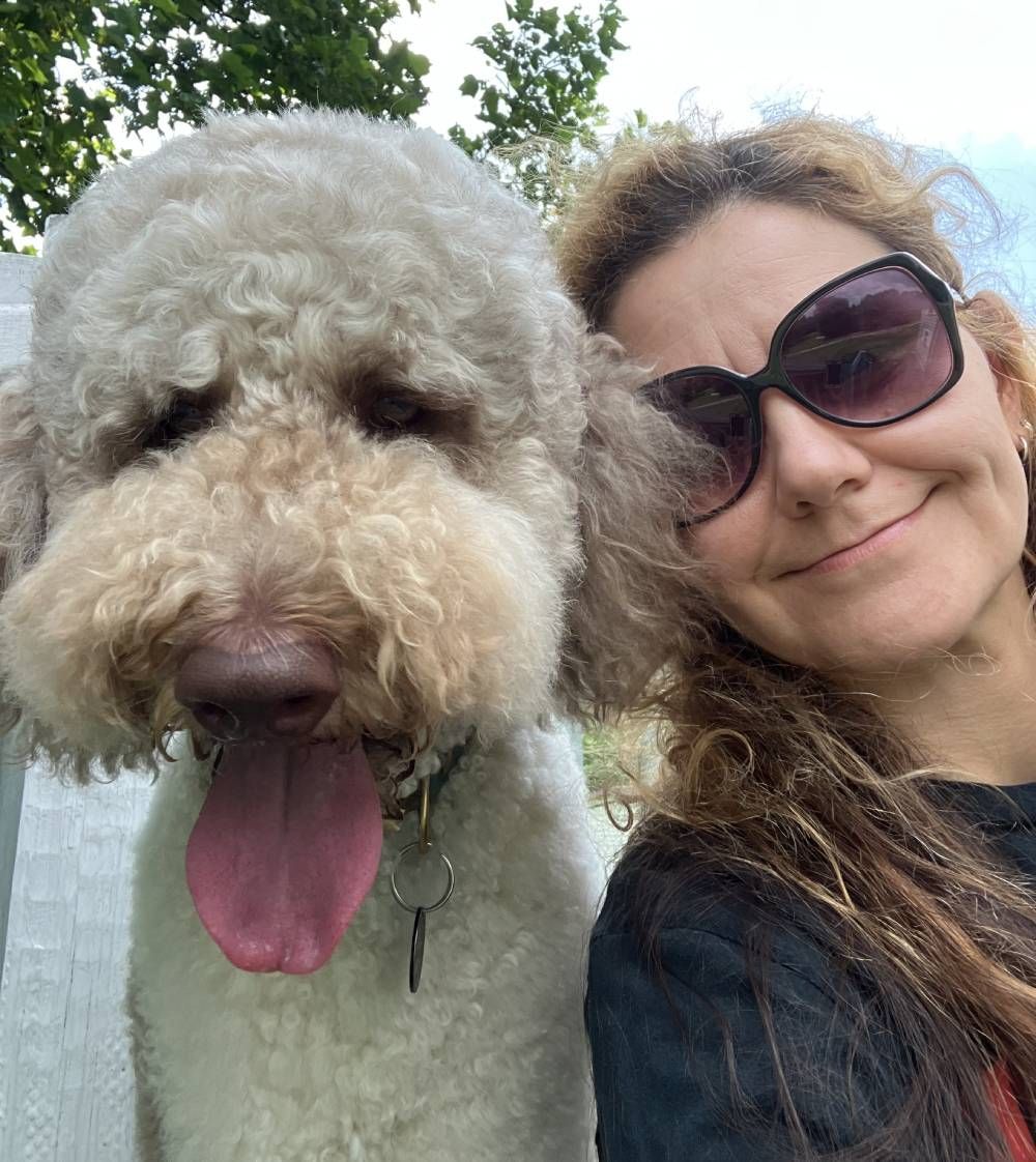A person with wavy hair and sunglasses smiles next to a fluffy, cream-colored dog with its tongue out.