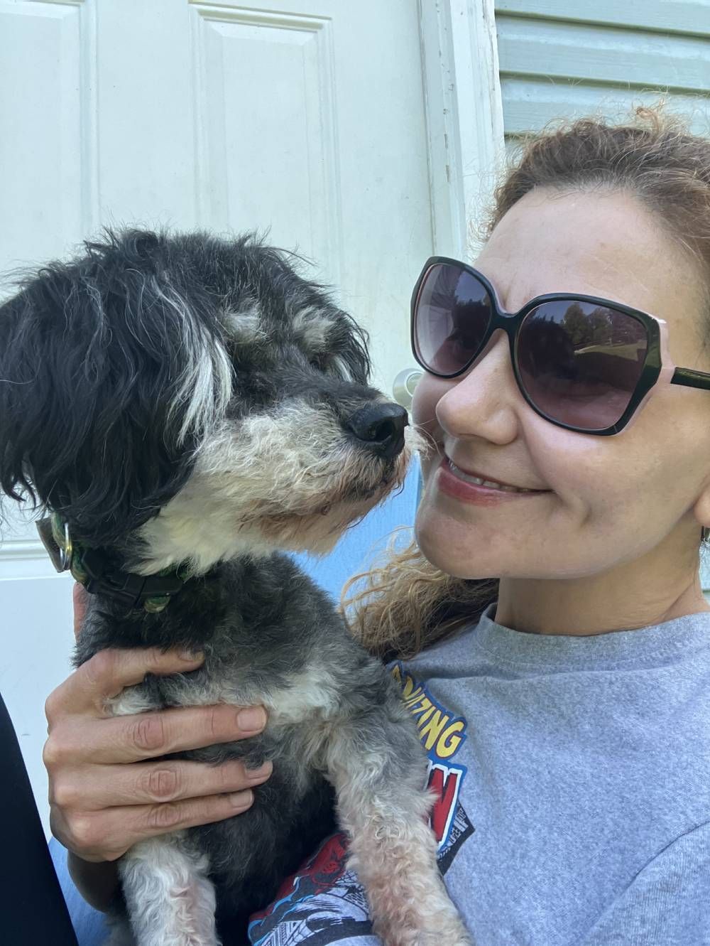 A person wearing sunglasses smiles while holding a small black and white dog in front of a white door.