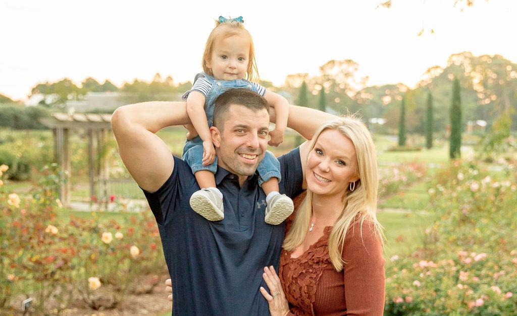 A man and woman are holding a baby on their shoulders in a park.
