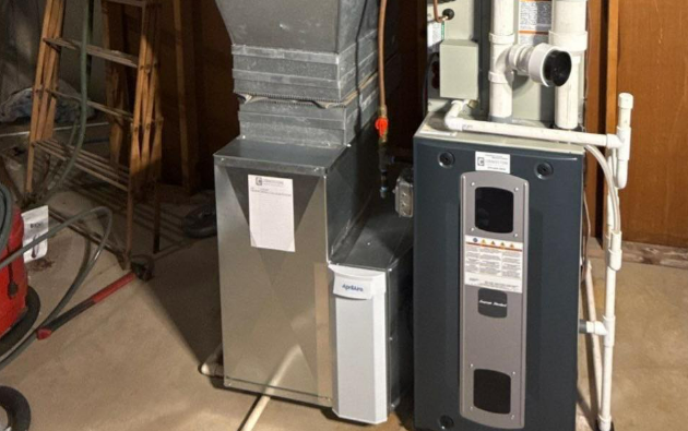 Furnace and water heater side-by-side in a basement. Metal and gray appliances against wood paneling.
