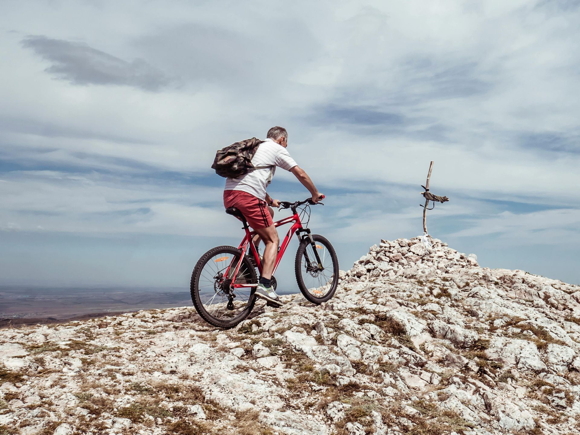 A person riding a mountain bike uphill toward a rocky peak marked by a cross under a cloudy sky.