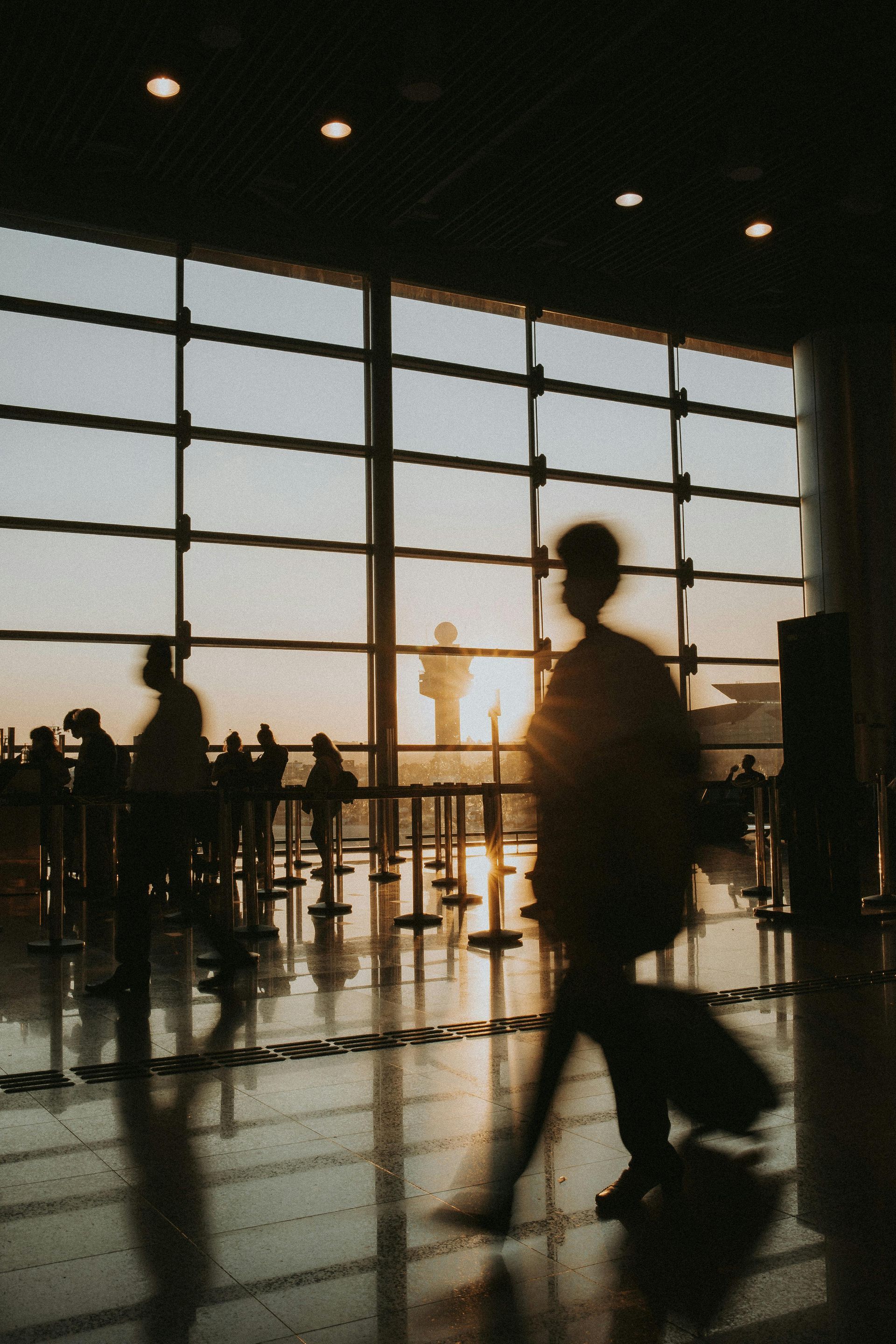 Silhouettes of travelers with luggage walking past a large airport terminal window at sunset.