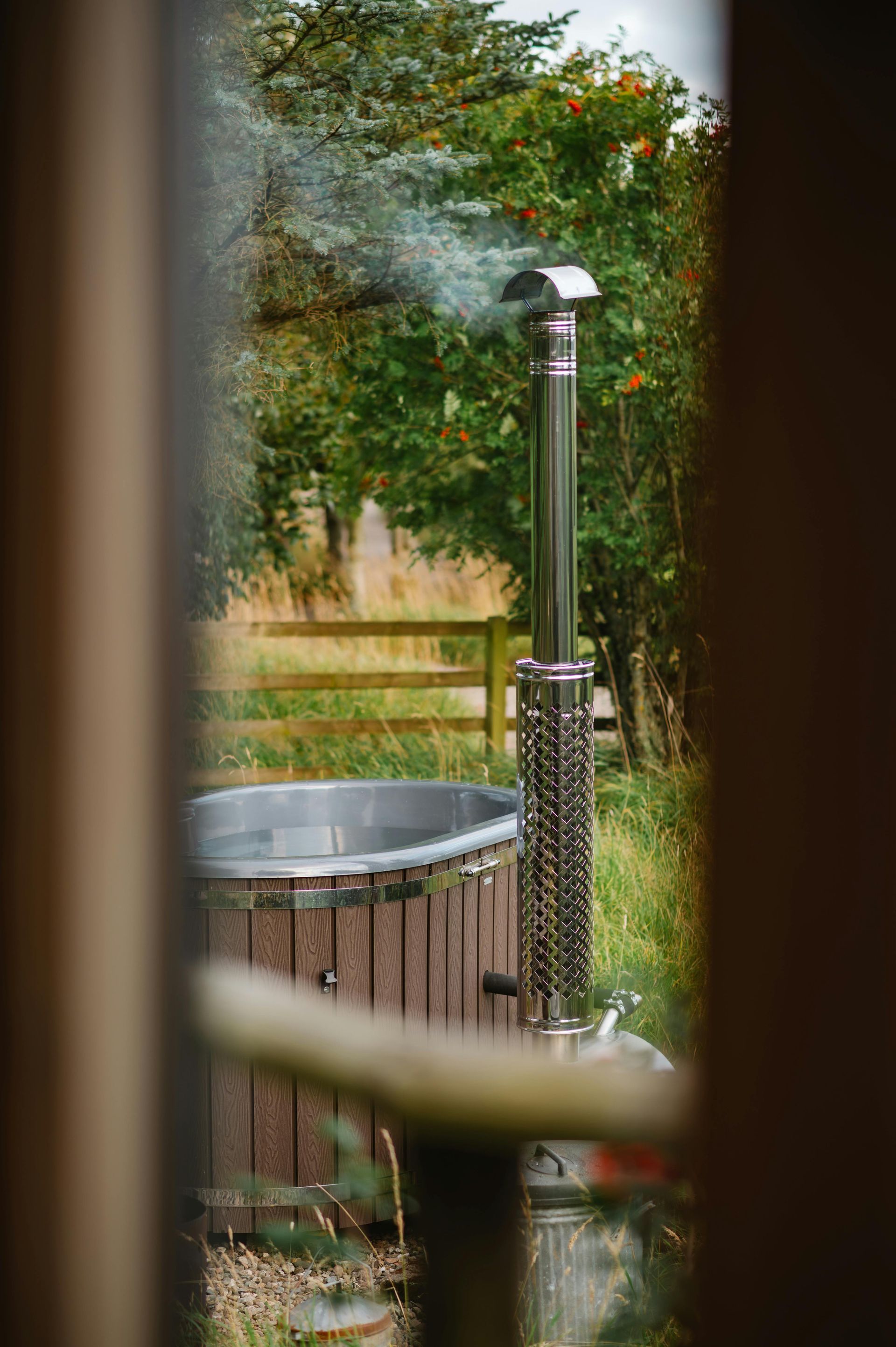 A wooden hot tub with a steaming metal chimney stands in a lush garden, viewed through a framed opening.