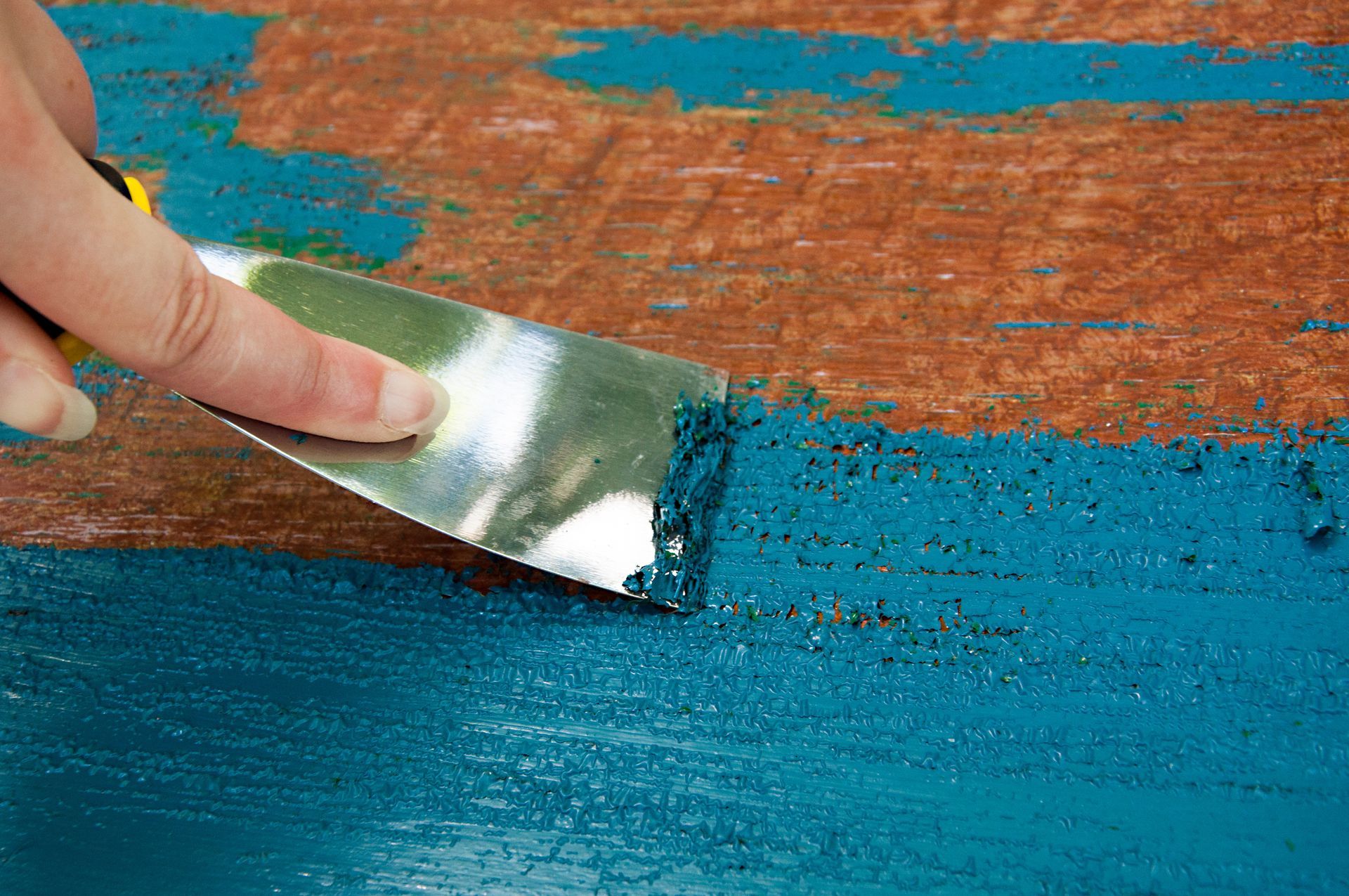 A person is using a spatula to remove paint from a wooden surface.