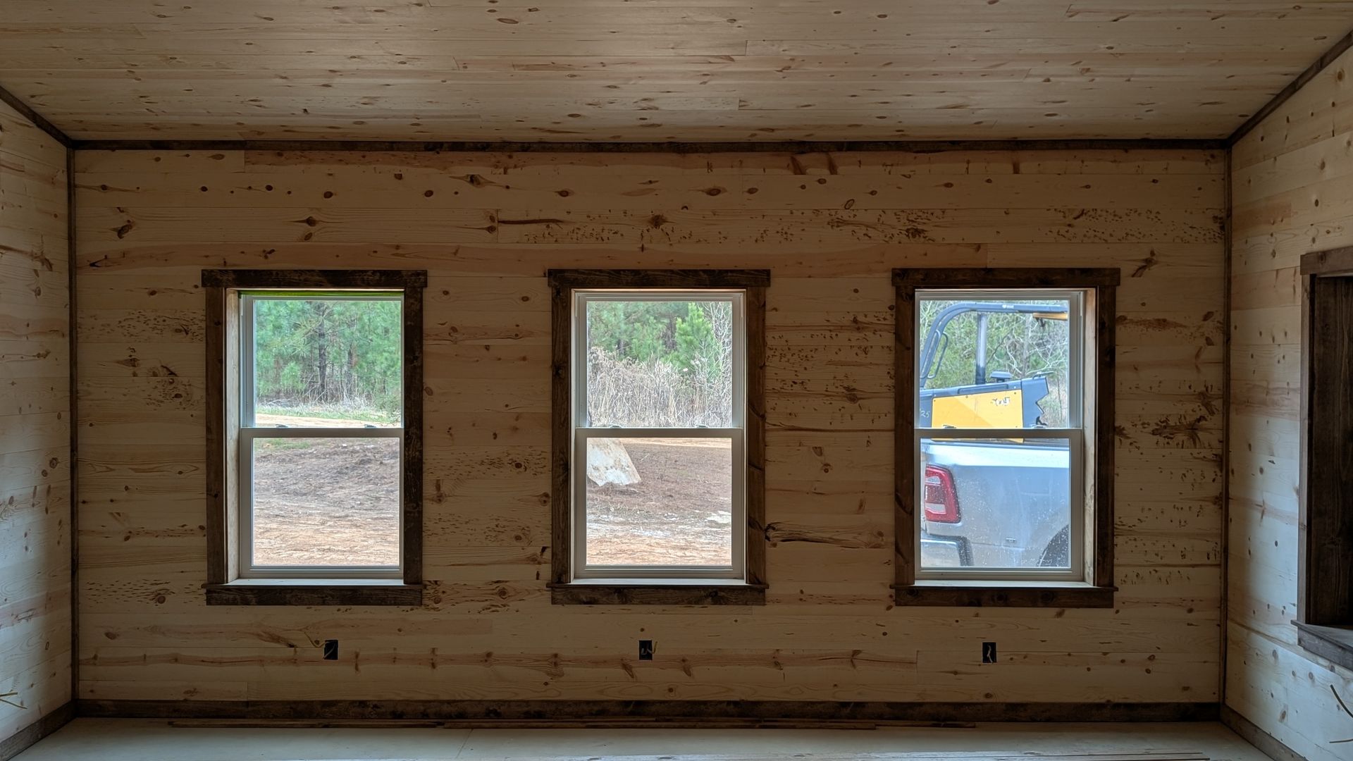 Interior view of a room under construction with three windows. Wood paneling covers walls and ceiling; windows look out to trees and a vehicle.