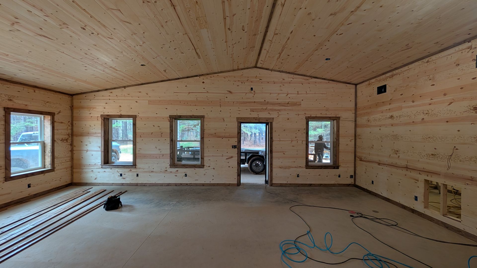 Interior of a room under construction, with wood-paneled walls and ceiling, windows, and an open doorway.