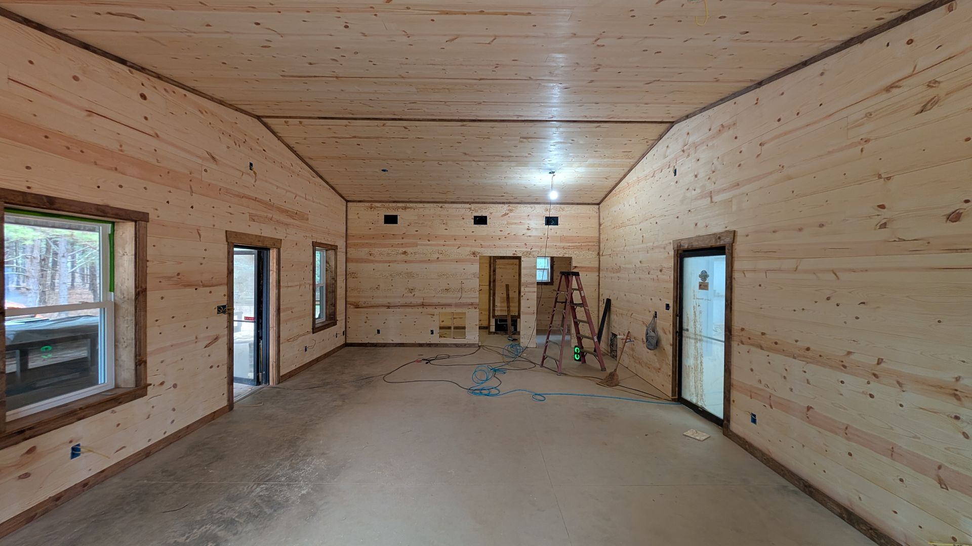Interior of a room under construction, with wood-paneled walls and ceiling, concrete floor, windows, and doorways.
