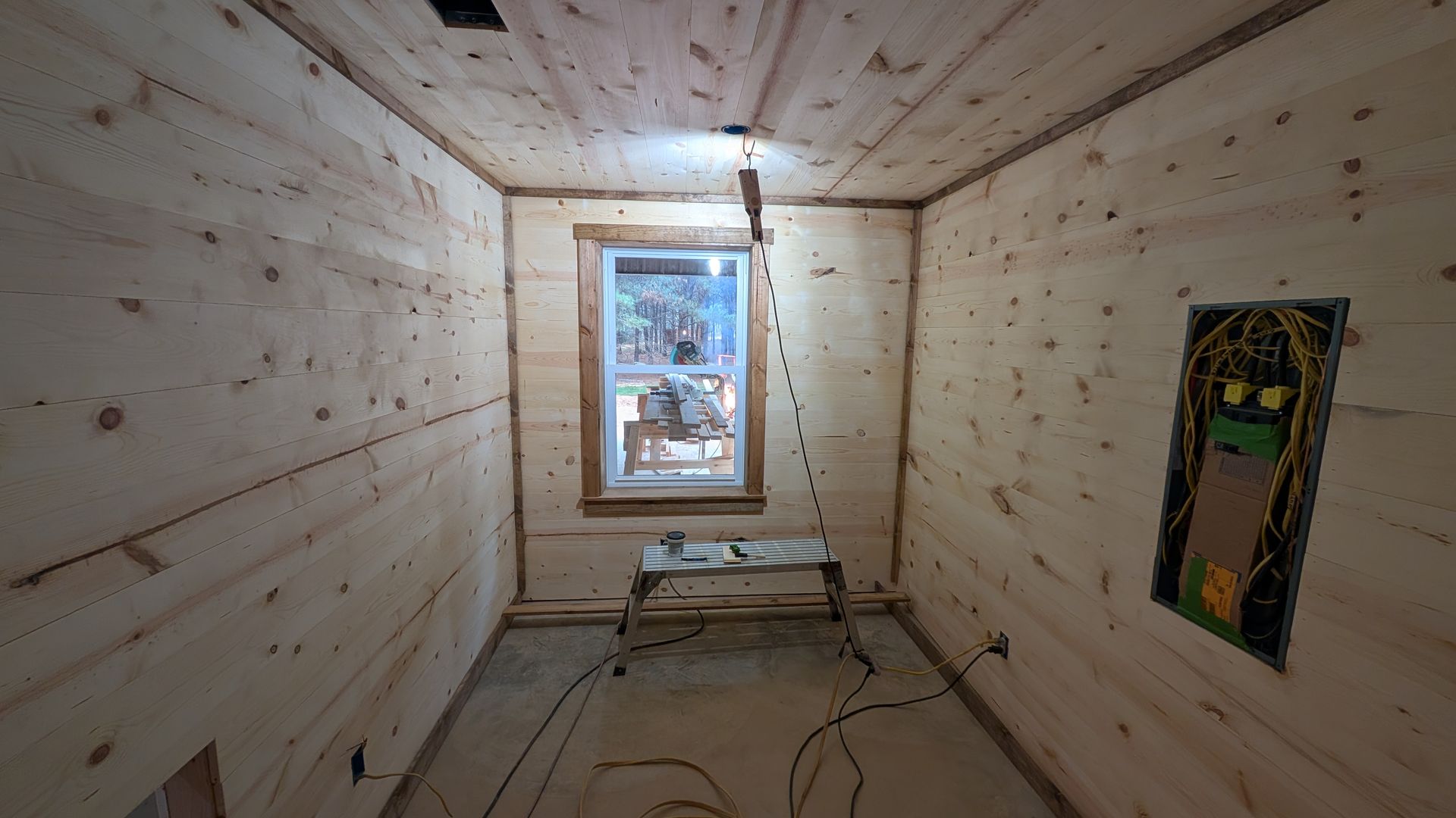 Interior of a room with wooden walls and ceiling. A window is in the center, a circuit box is on the right wall.