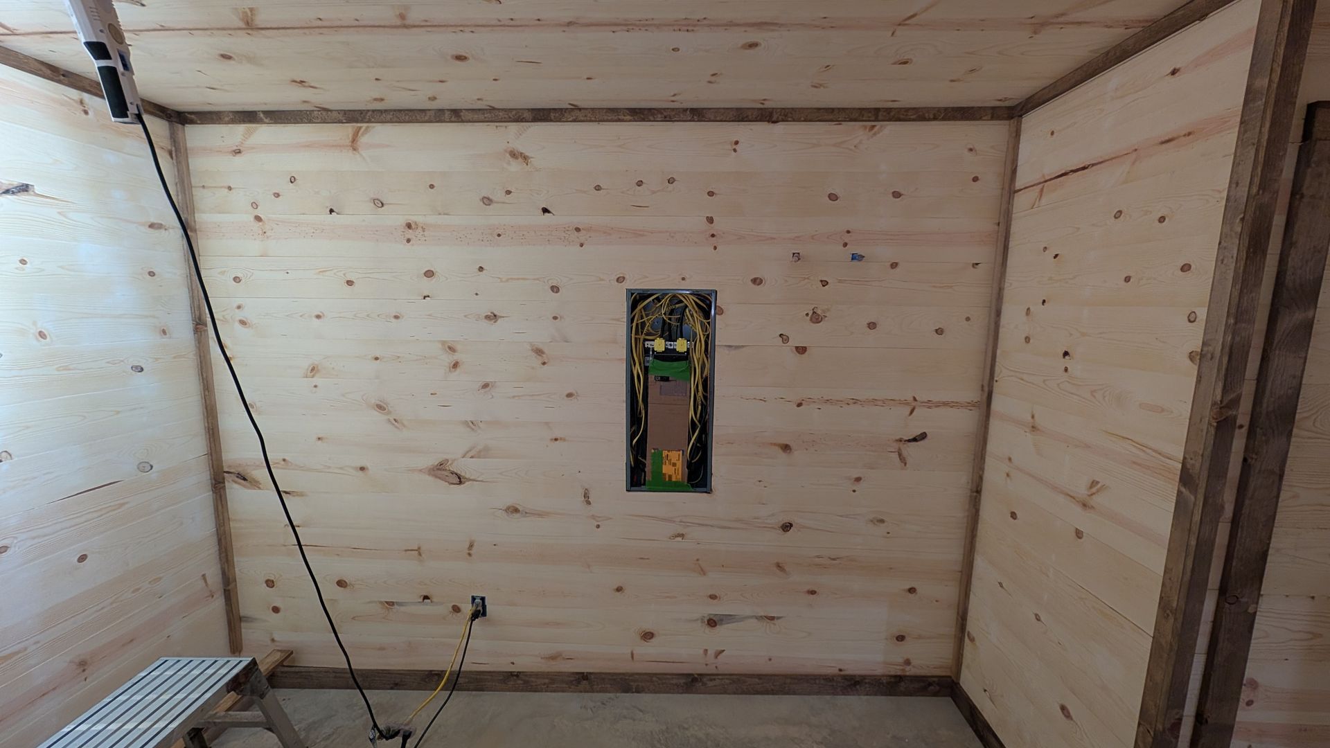 Interior of a wood-paneled room with electrical panel in the center. A black cord hangs from the ceiling.