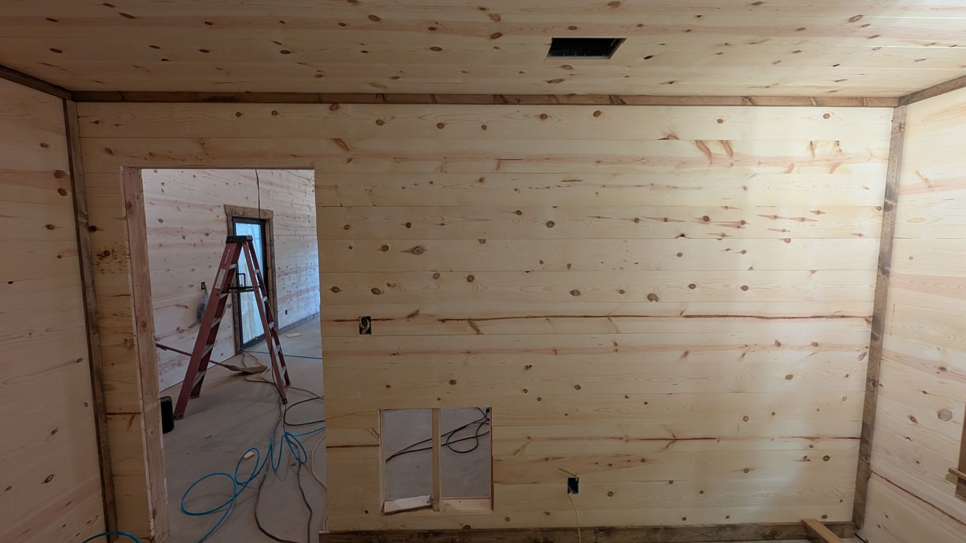 Interior of a room with unfinished wood paneling on walls and ceiling. Doorway and small window opening visible.