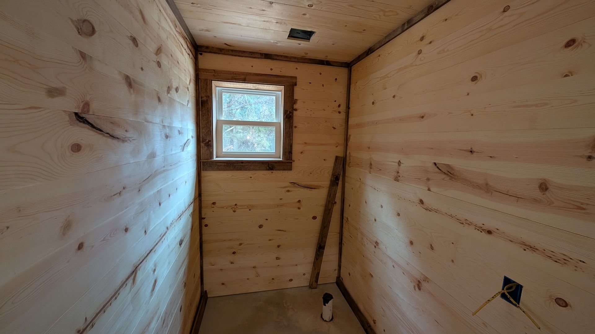 Interior of a small room with unfinished wooden walls, window, and electrical outlet.