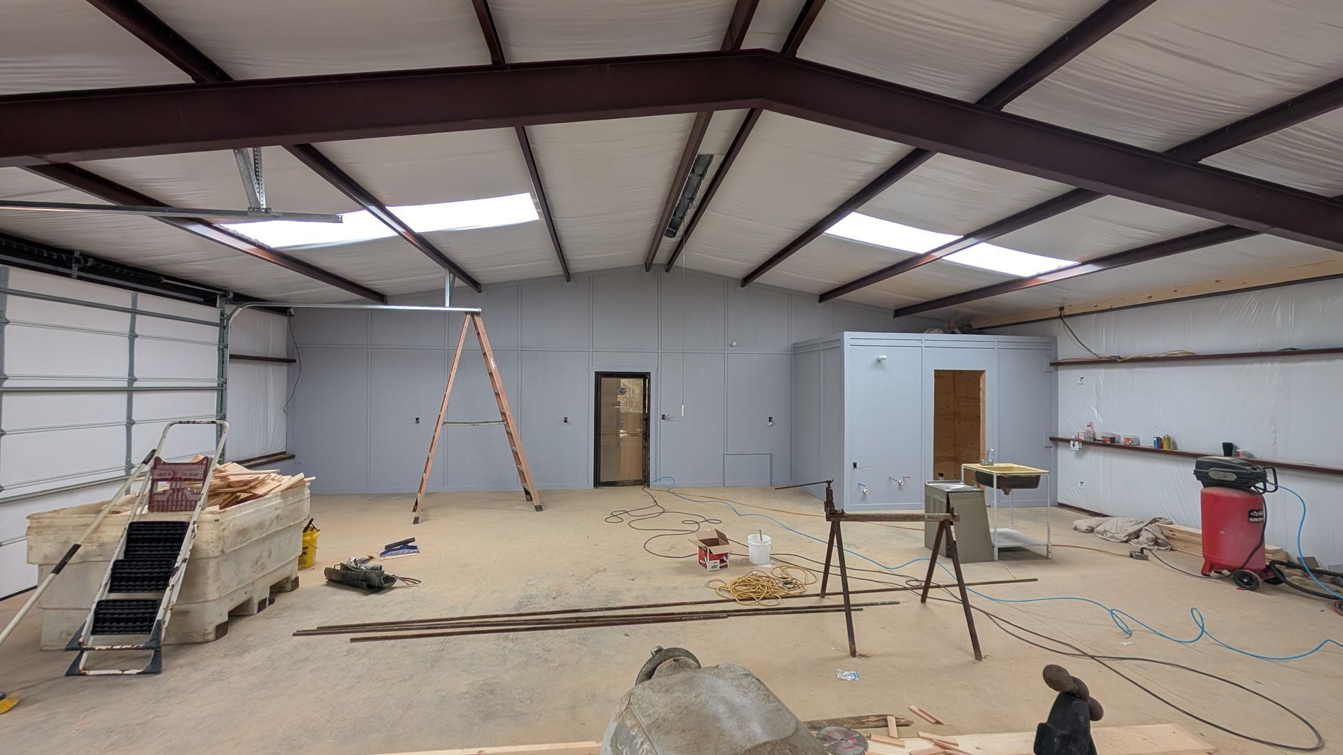 Interior of a large unfinished metal building. Light-colored walls, exposed steel beams, skylights, construction debris.