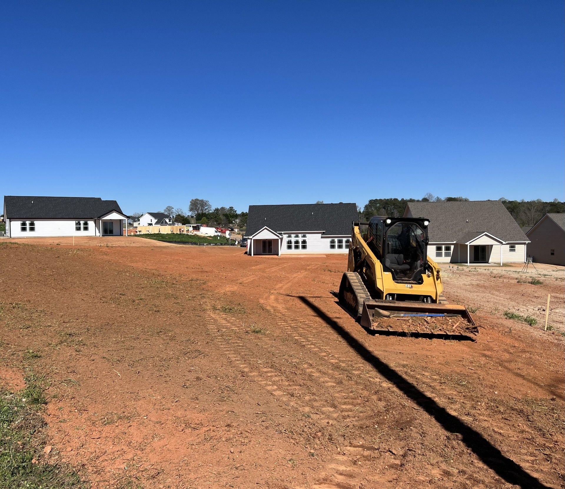 A yellow skid steer tractor on a construction site with houses in the background under a blue sky.