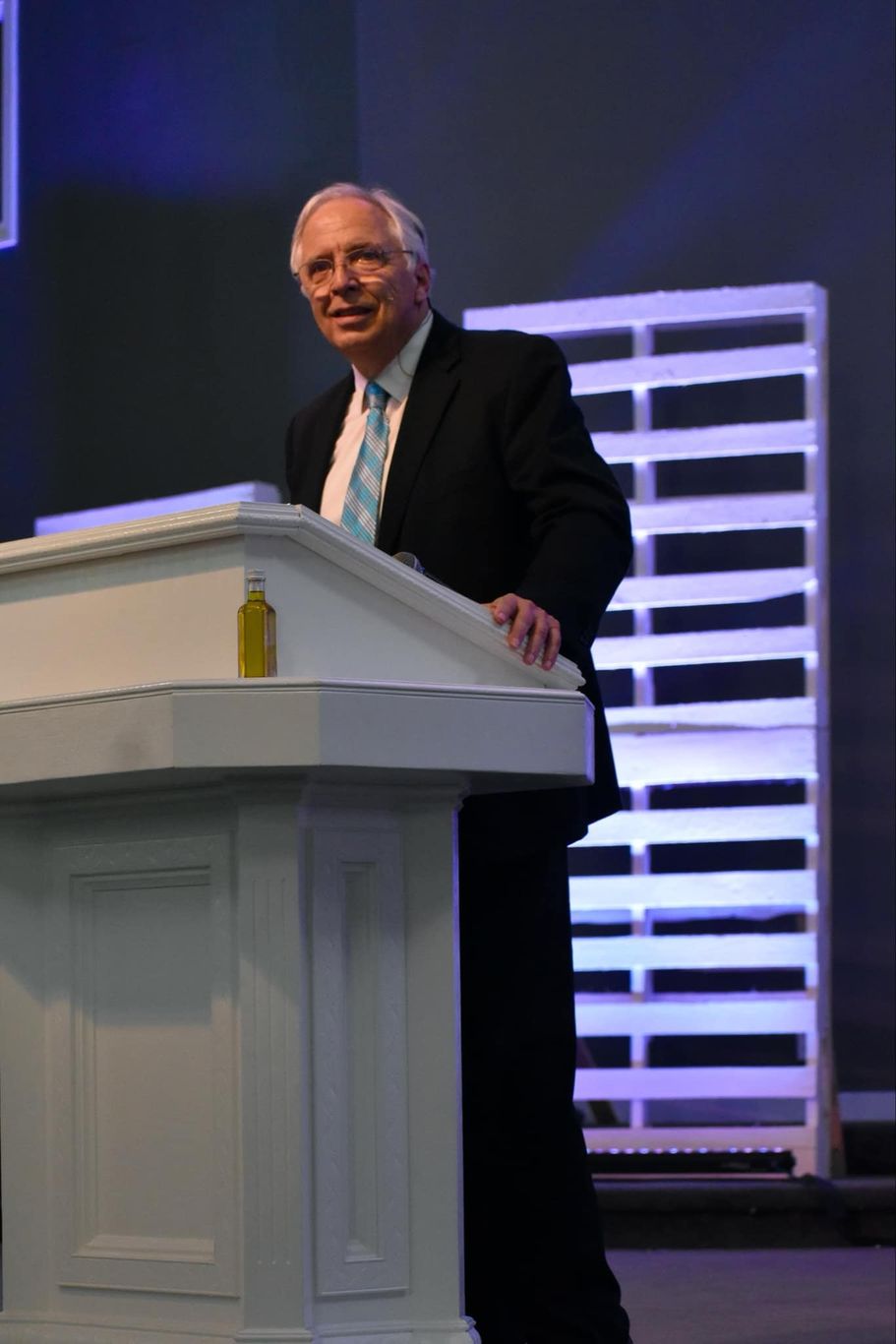 Man in suit, standing at a podium, speaking. Stage with blue and white lights.
