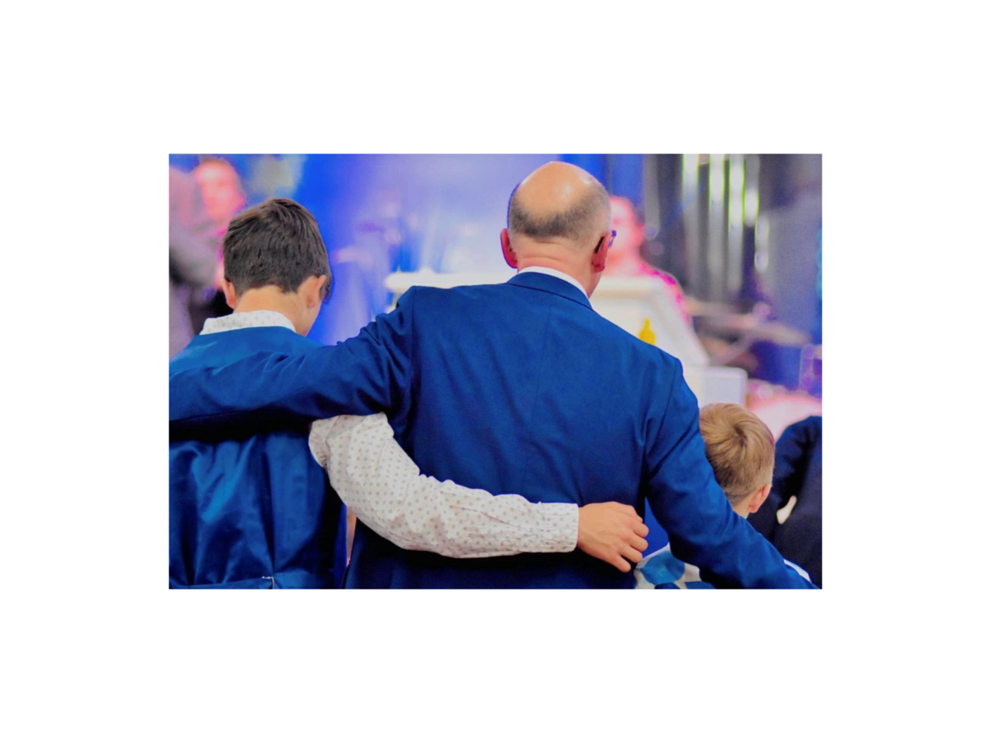 Man in blue blazer with arm around a younger person's shoulders. Other people in background.