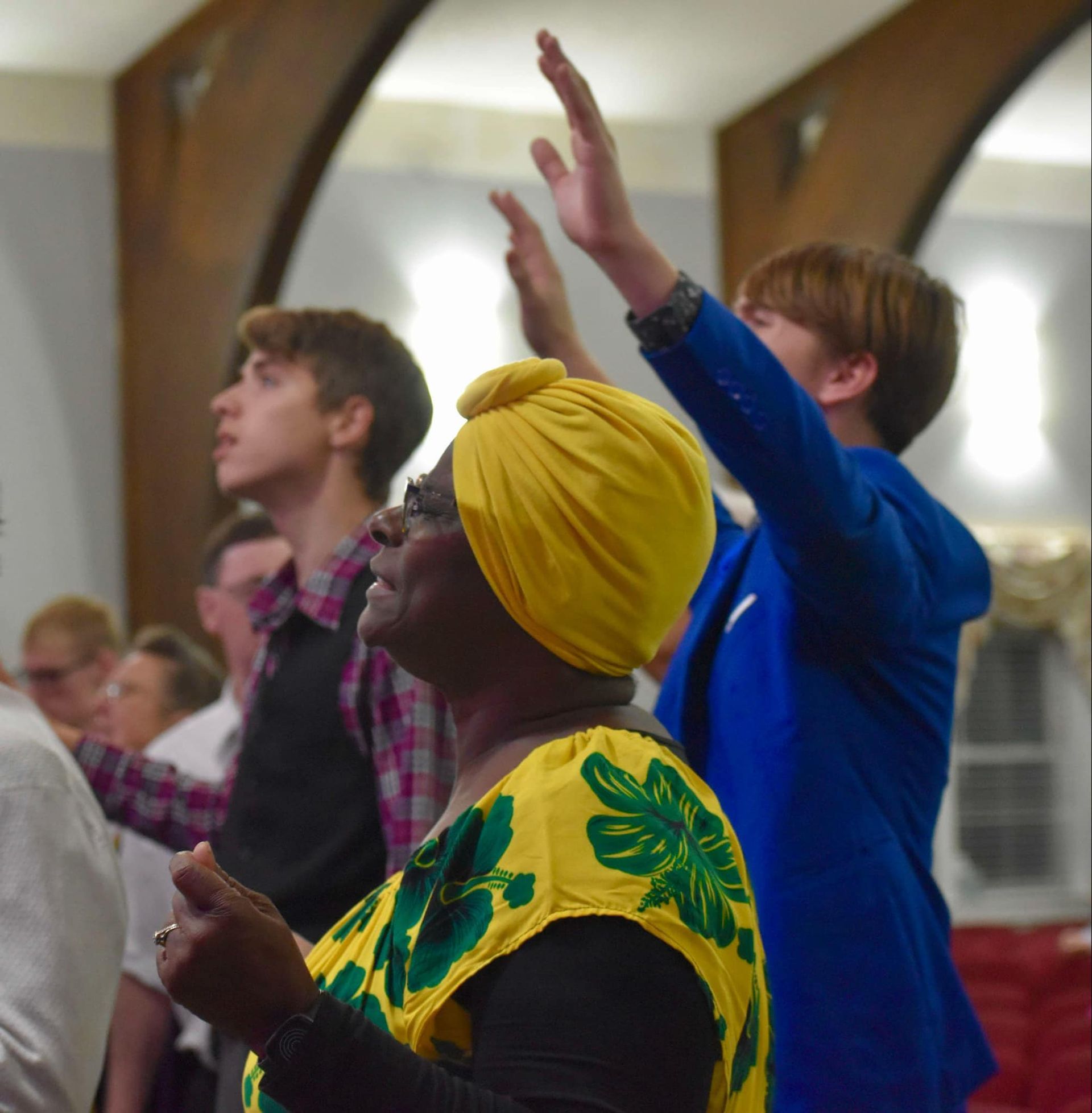 Woman with yellow turban and shirt sings, hands raised, with others in a church.