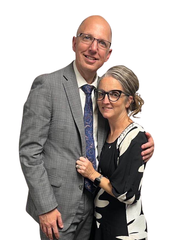 Man and woman in formal attire, smiling, embracing. Man bald, in suit. Woman has glasses, patterned dress. White background.