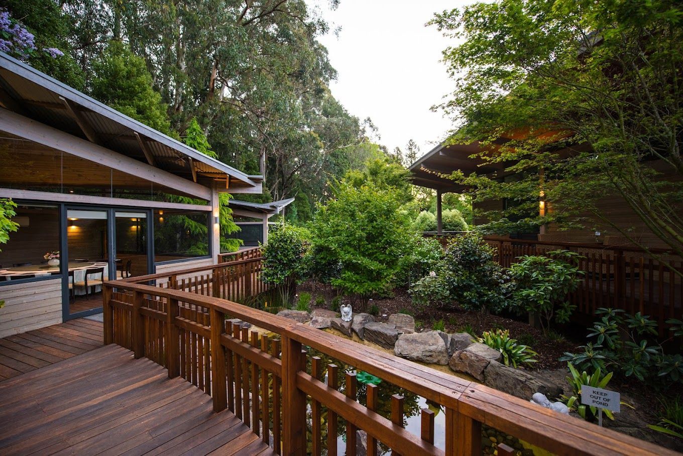 Wooden walkways and buildings surrounded by lush greenery and a small pond.