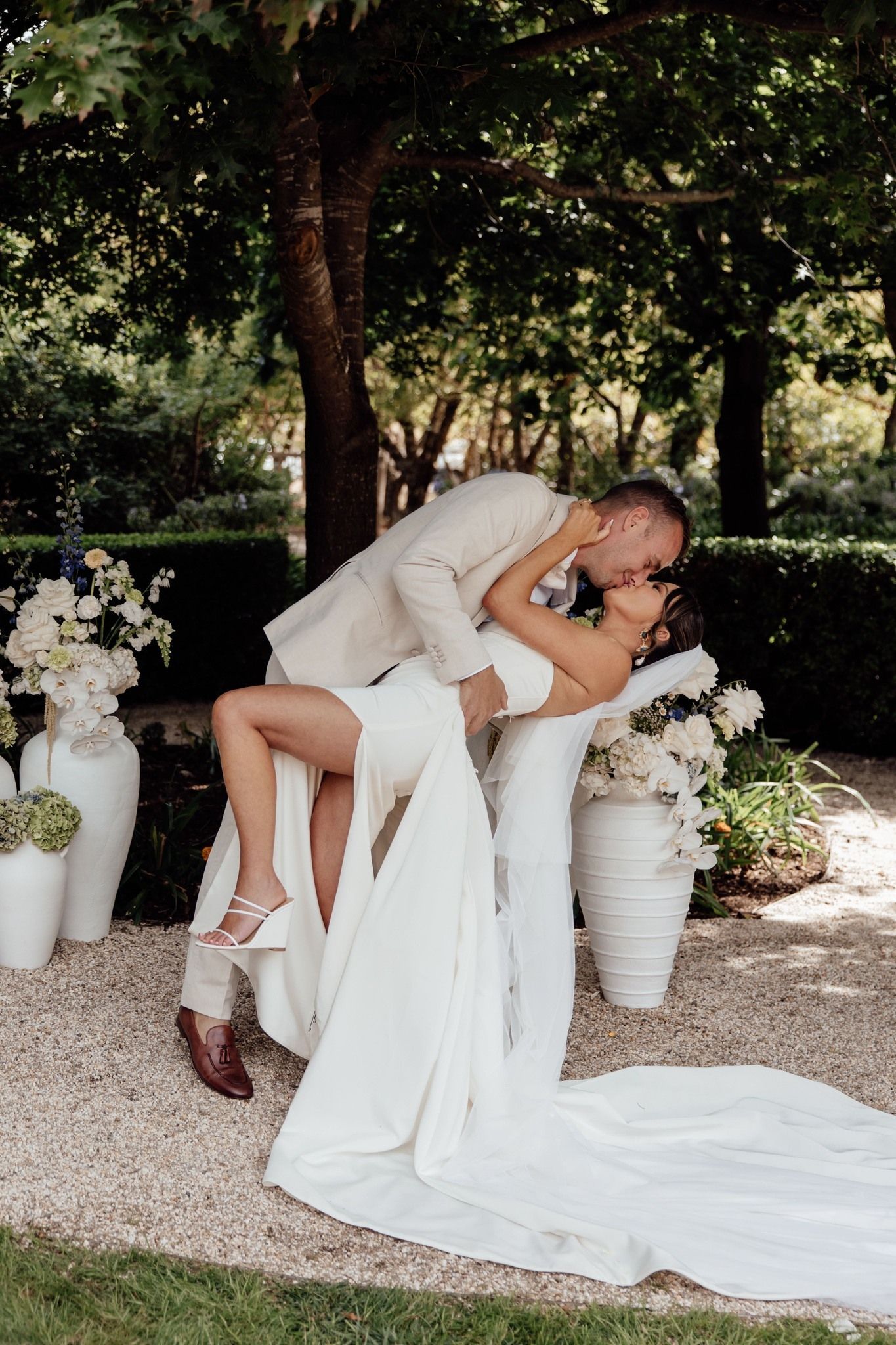 A bride and groom are kissing in a garden . the bride is wearing a long white dress.