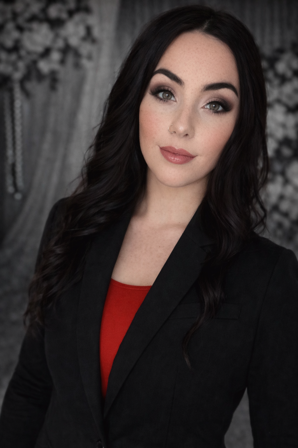 Woman in black blazer, red top, looking at the camera; blurred floral backdrop.