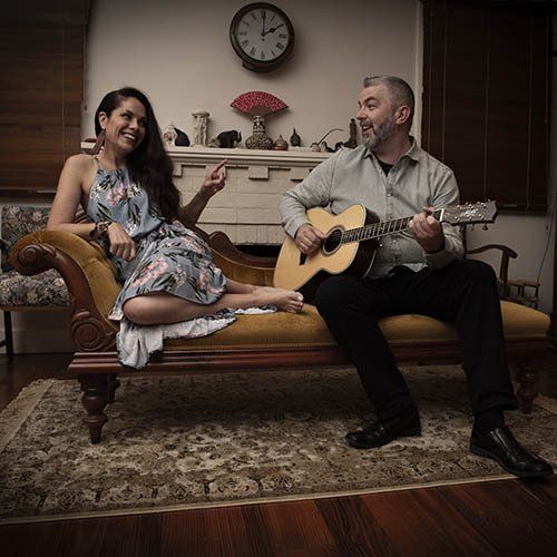 A man is playing a guitar while a woman sits on a couch