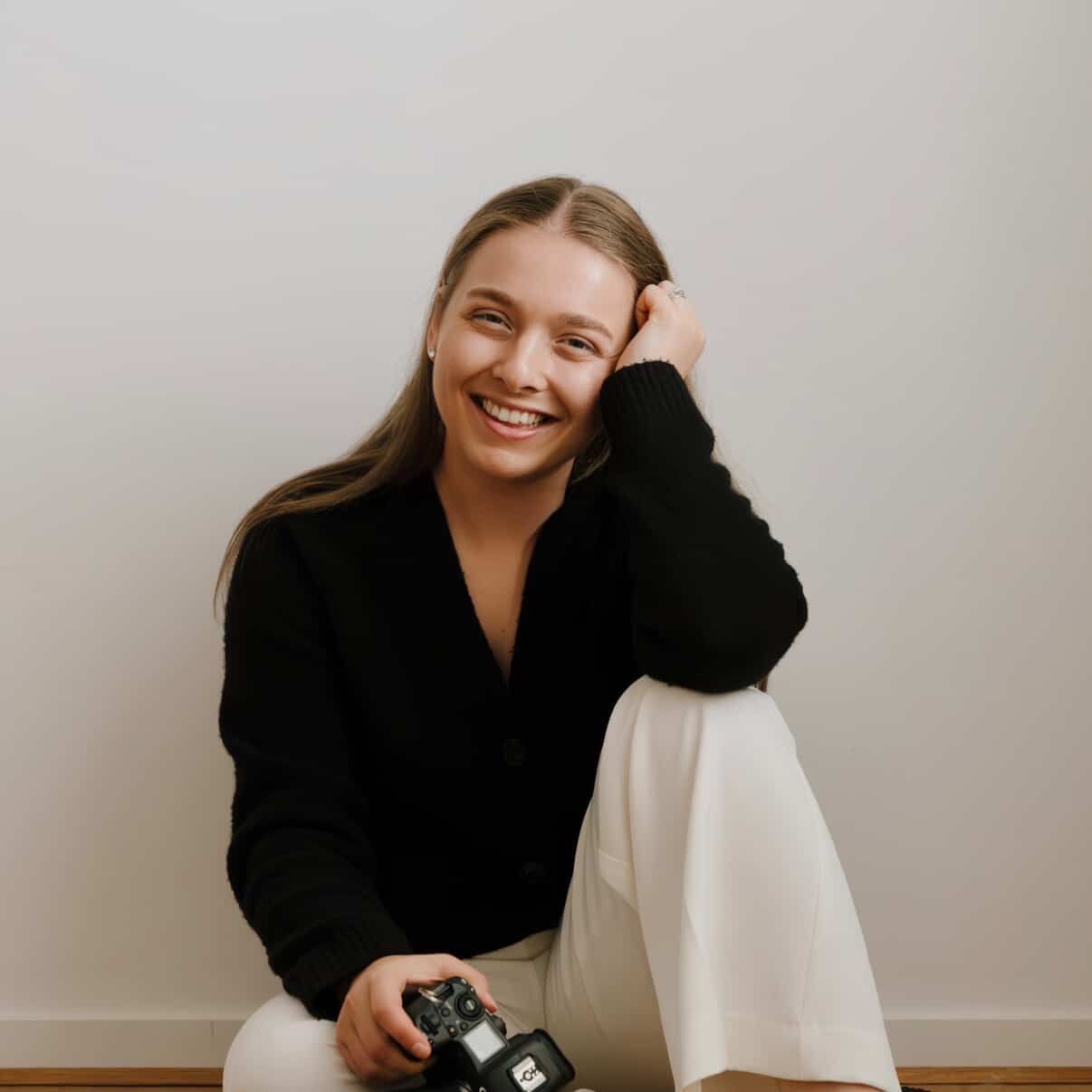 Woman smiling, holding a camera, sitting cross-legged in a bright room, wearing black sweater and white pants.