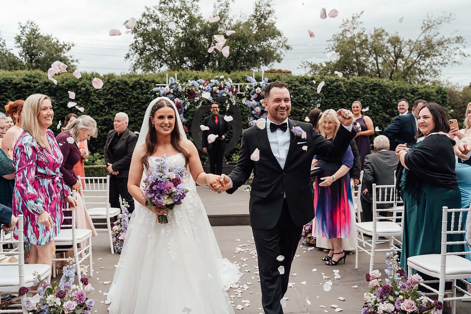 Newlyweds exiting wedding ceremony, holding hands, with guests cheering and flower petals falling.