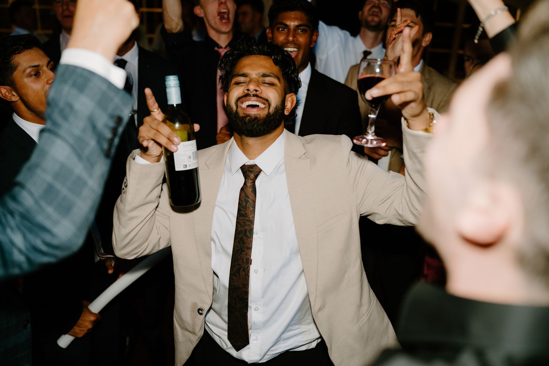 A man in a suit and tie is dancing in a crowd while holding a bottle of wine.