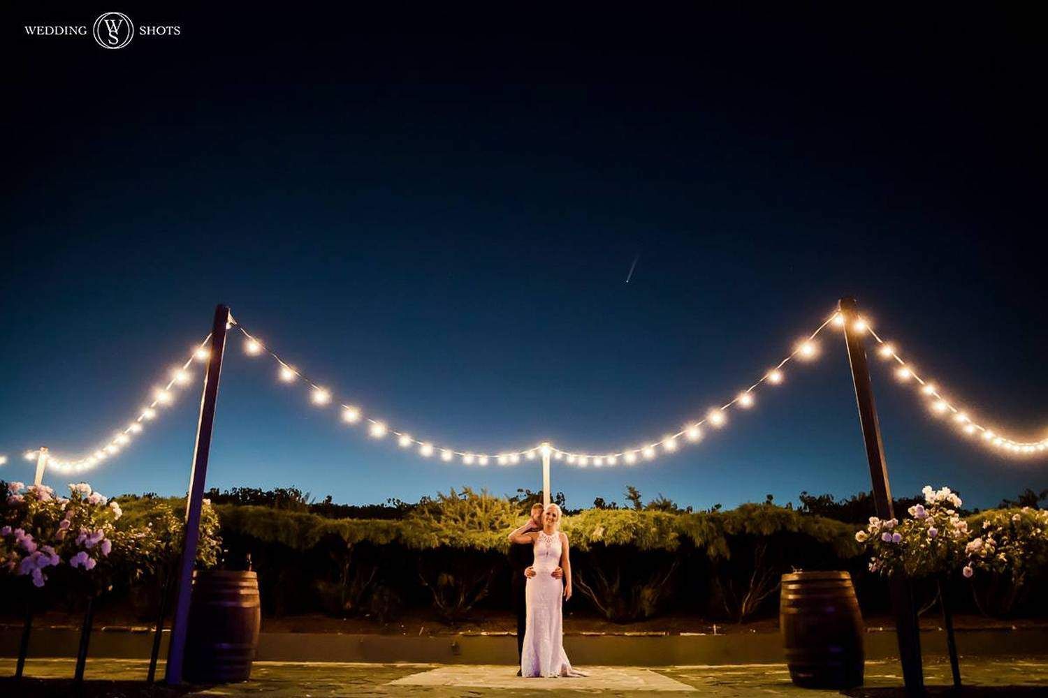 A bride and groom are standing under a string of lights at night.