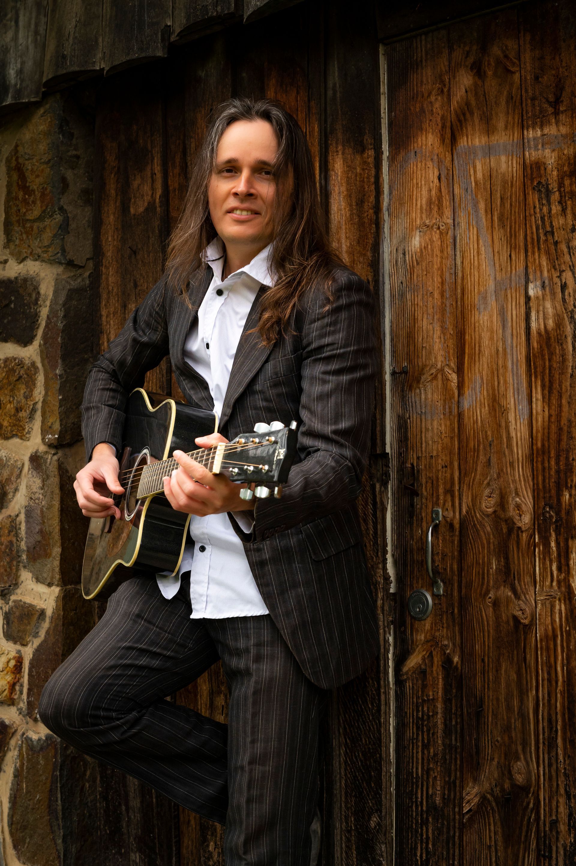 A man in a suit is holding a guitar and leaning against a wooden door.