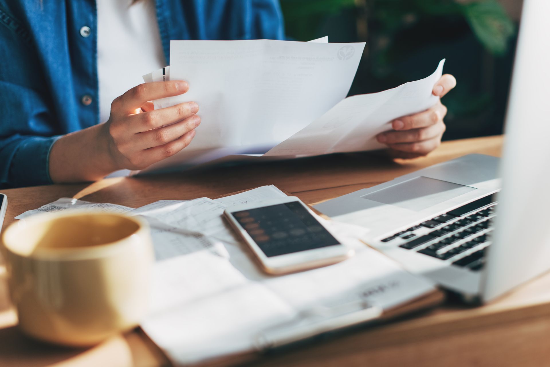 Hands holding a black spiral-bound bookkeeping notebook on a desk, with other papers and a pen nearby.