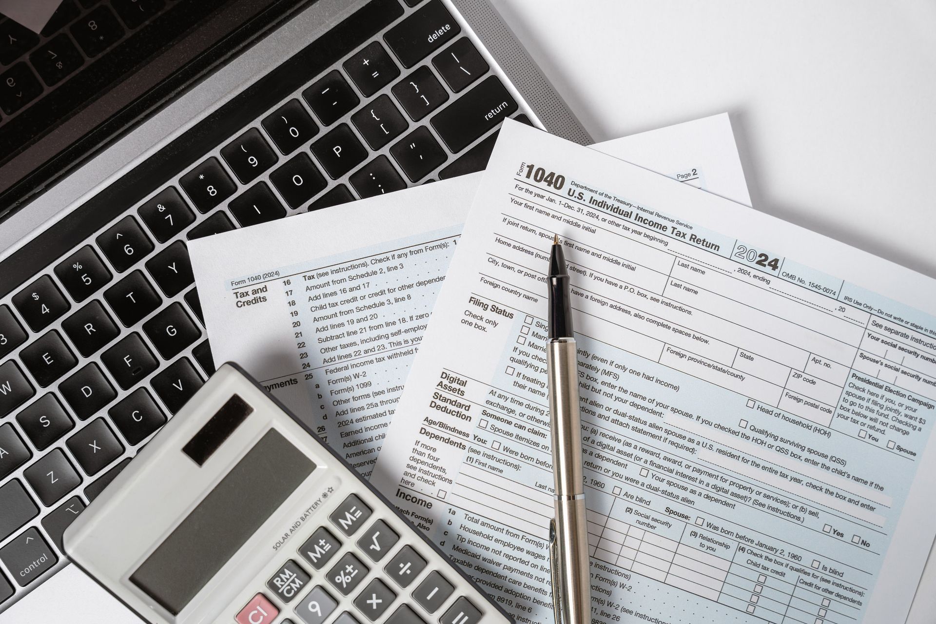 Laptop, tax forms, calculator, and pen on a white surface, representing tax preparation.