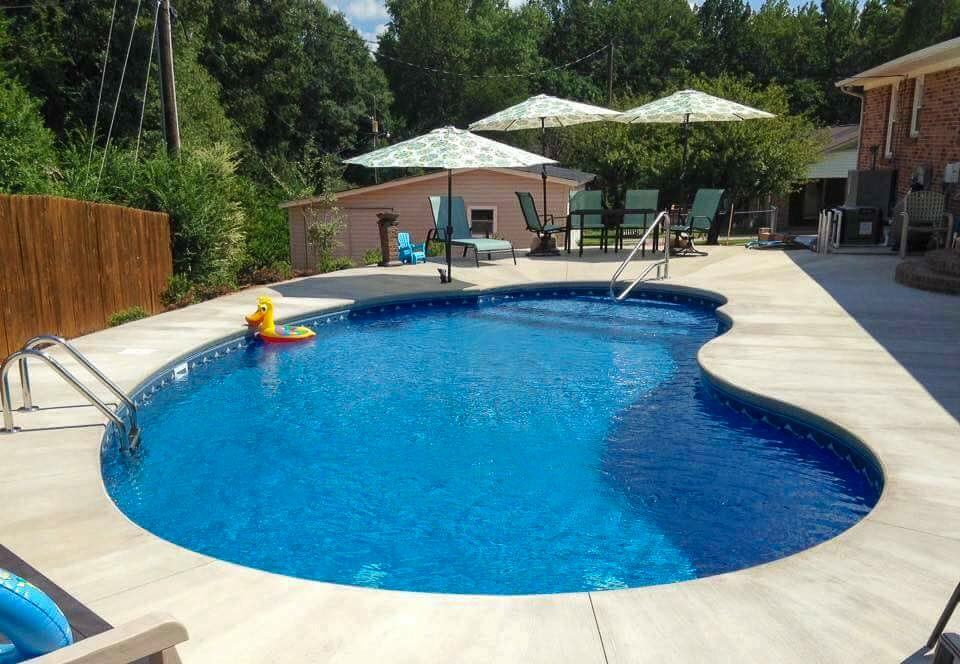A backyard pool with blue water, surrounded by concrete and lounge chairs under umbrellas.