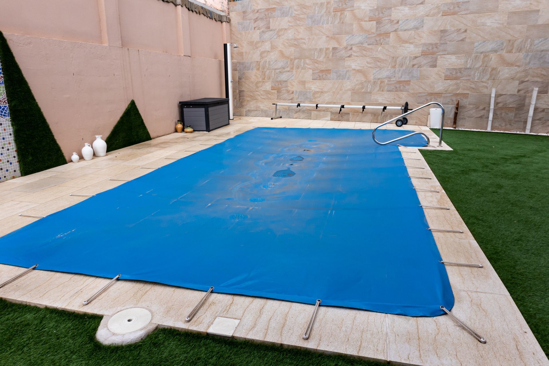 Backyard pool covered with a blue tarp, surrounded by a tile deck and green accents.