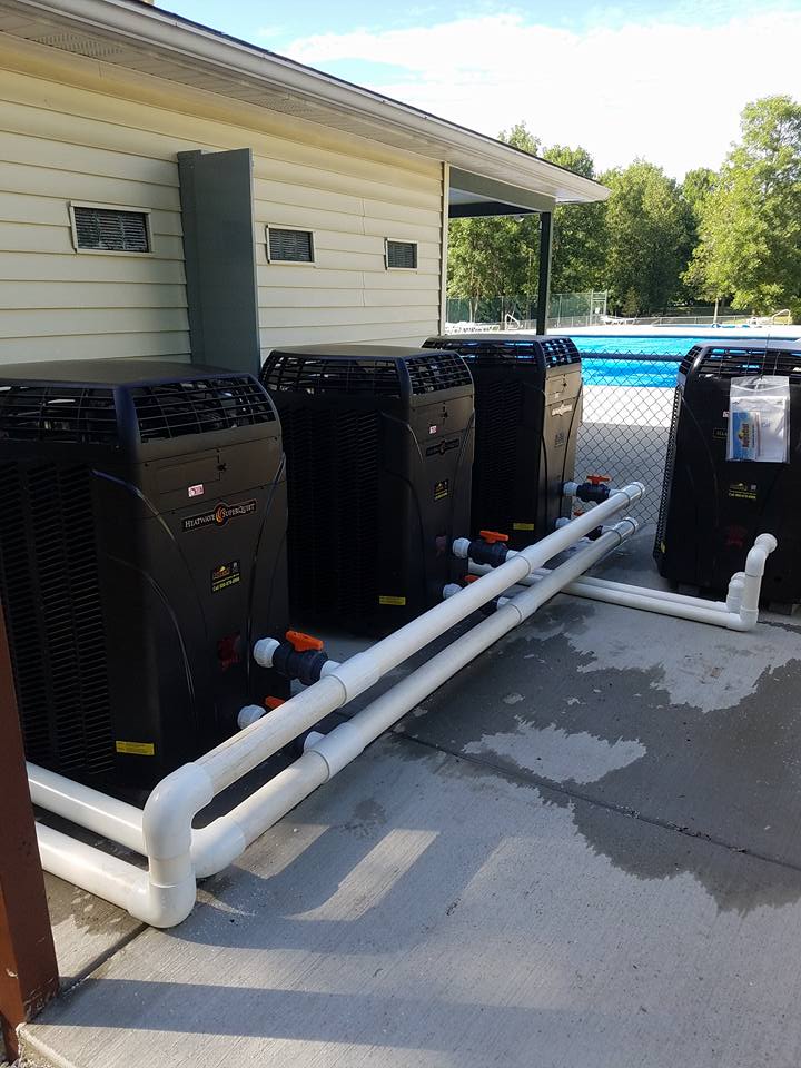 Four black pool heaters with white pipes, on a concrete pad next to a pool.