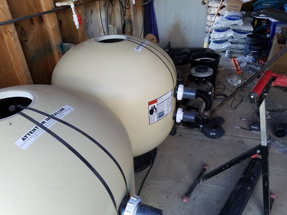 Two beige pool filter tanks with piping, fittings, and tools in a workshop.