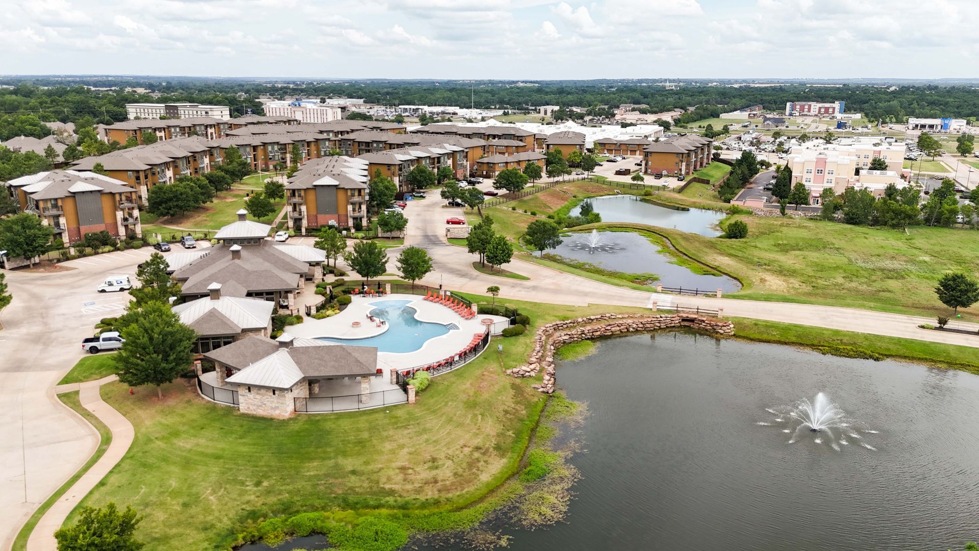 Aerial view of apartment complex with pool, pond, and fountain in green area under cloudy sky.