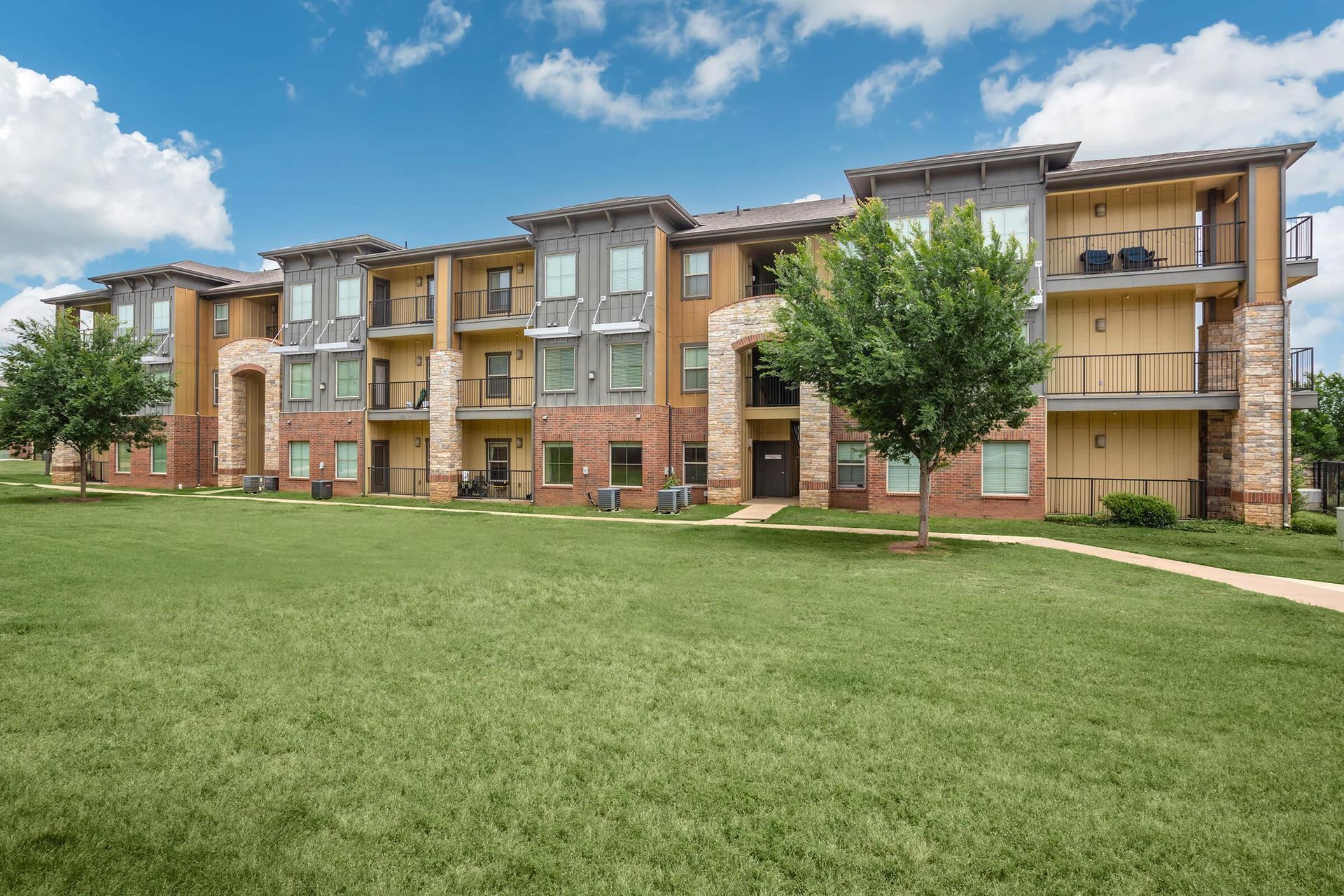 Apartment building with beige, red, and gray siding, green lawn, and blue sky.