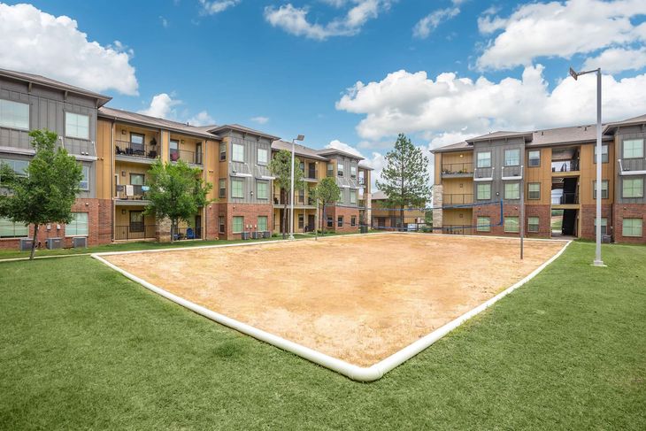 Sandy volleyball court surrounded by green grass, between two apartment buildings under a blue, cloudy sky.