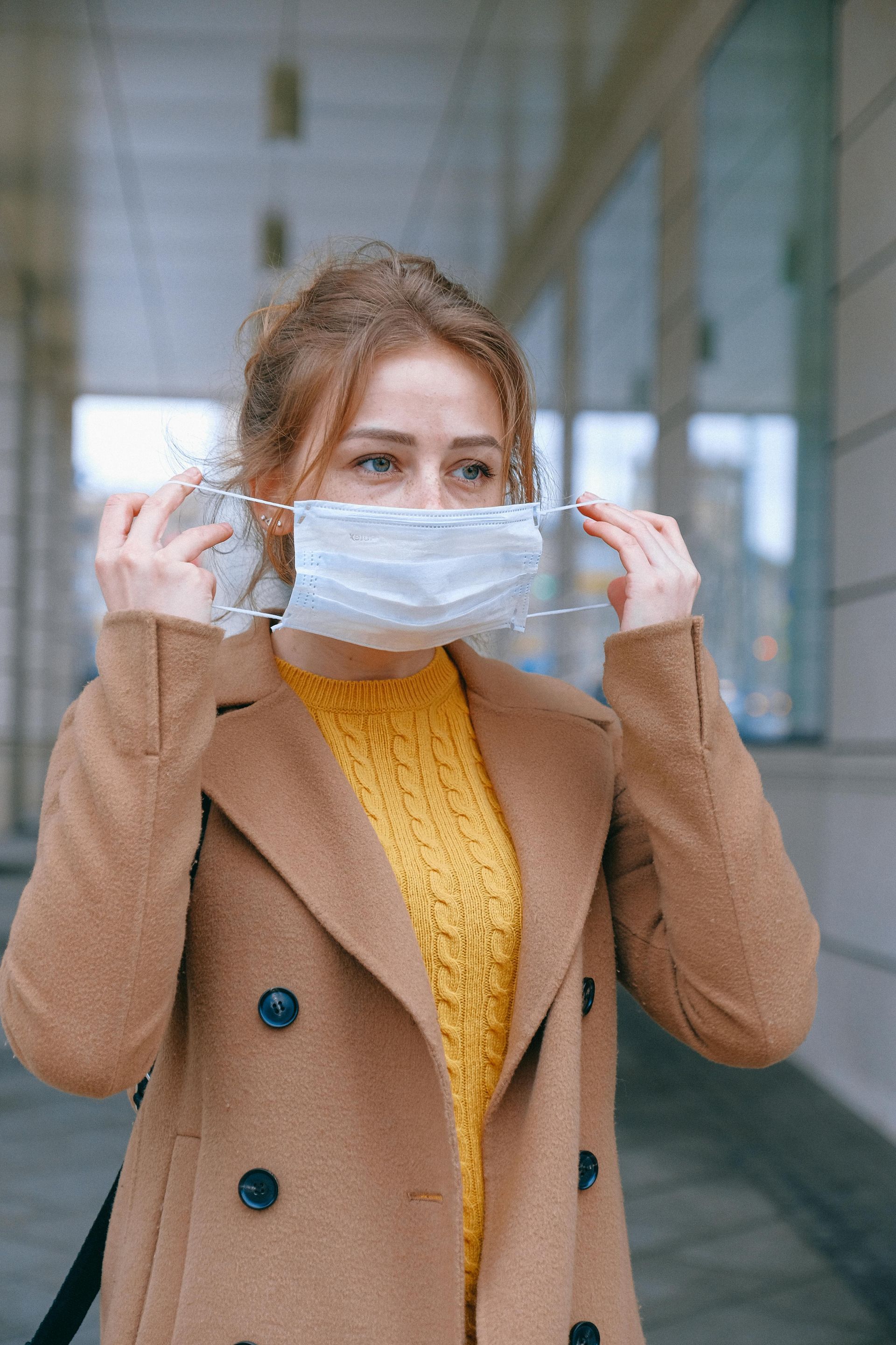 Woman in brown coat puts on a face mask outside, looking towards the side.