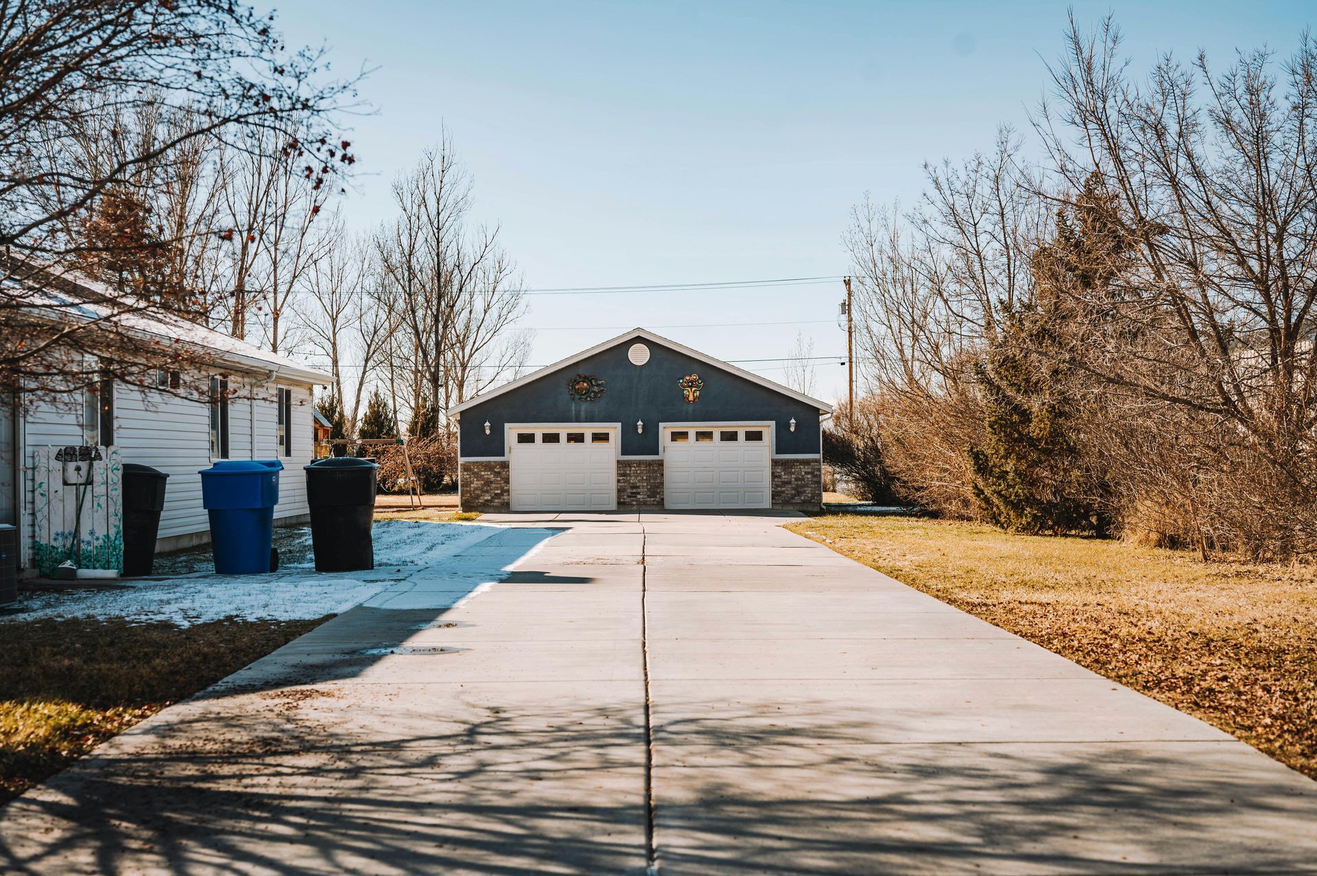 Suburban driveway leading to a two-car garage with leafless trees and blue trash bins on the left