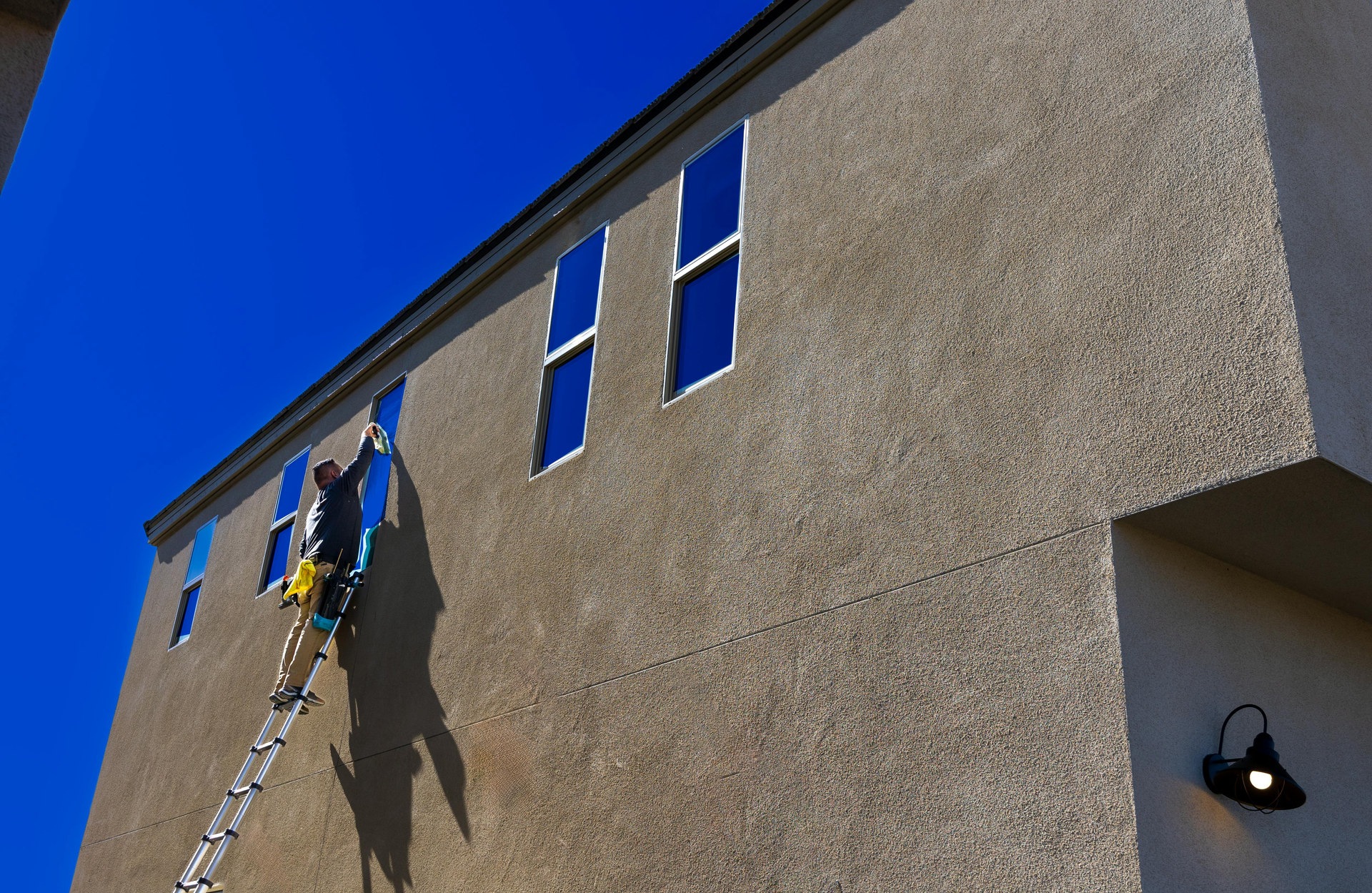 Worker on a ladder painting a tan stucco building under a bright blue sky
