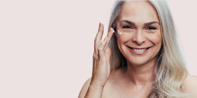 A woman is smiling while applying a cream to her face.