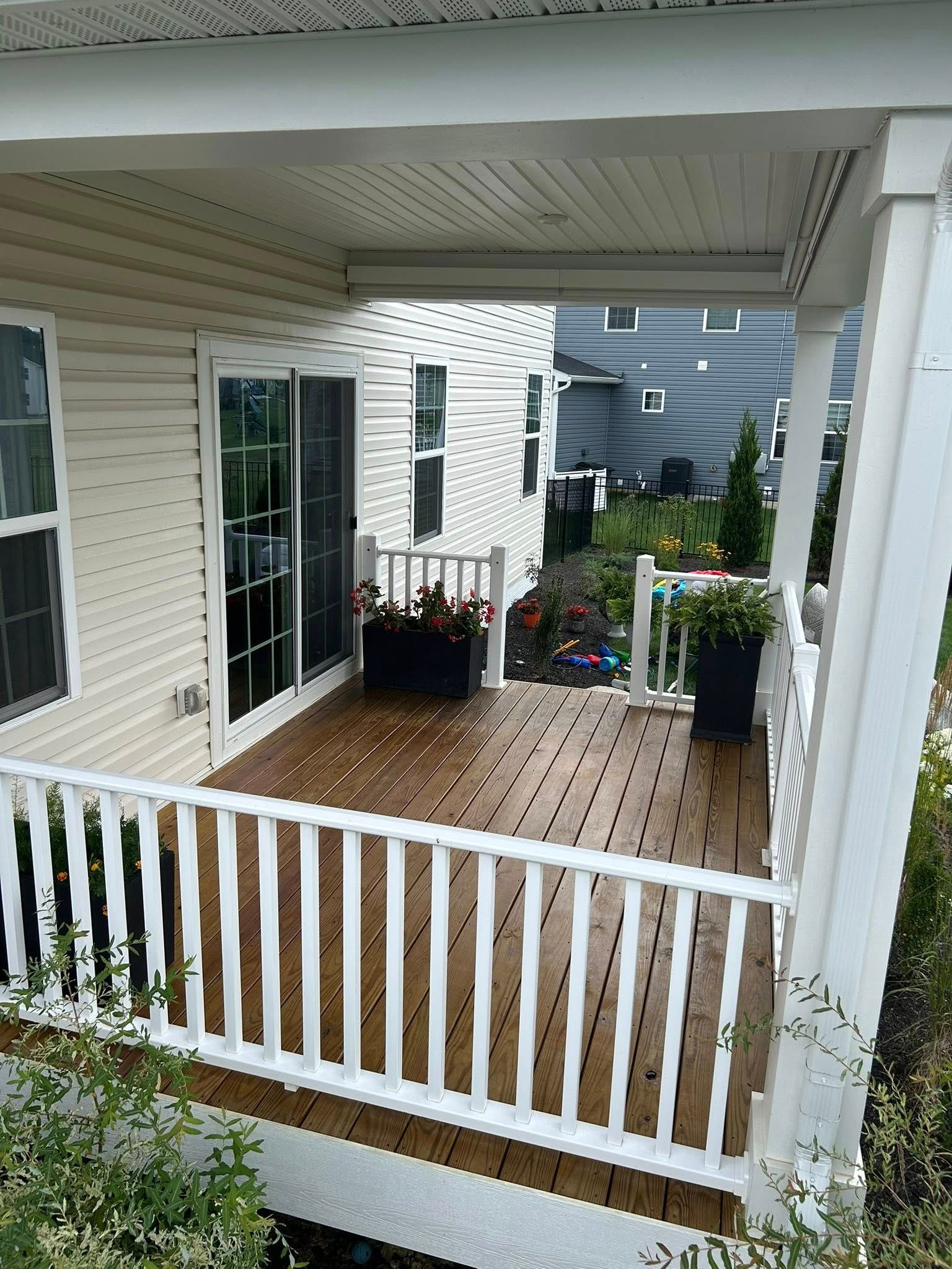 Wooden deck with white railing, overlooking a yard, with flowers and sliding glass door.