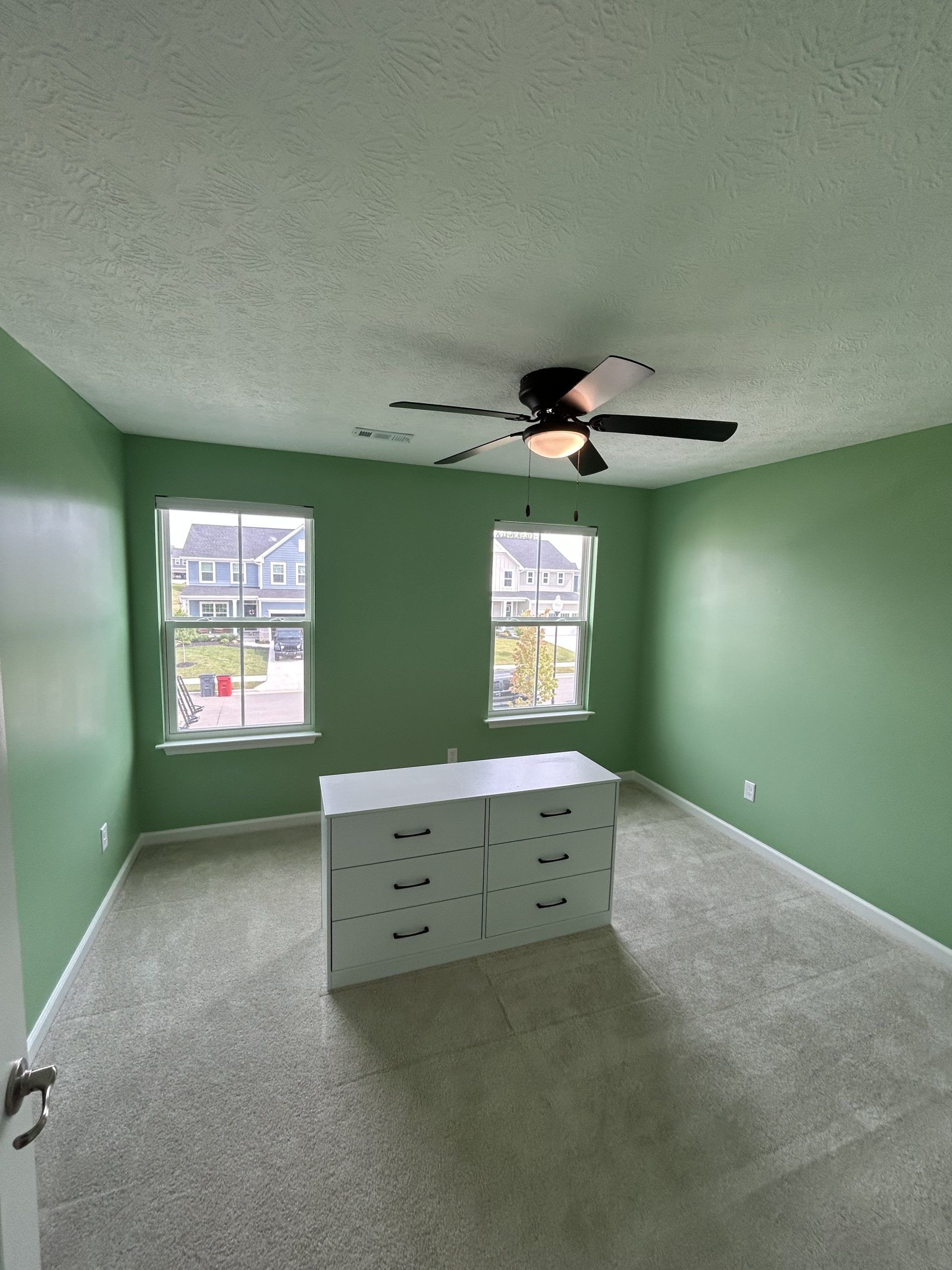 A bedroom with green walls, a white dresser, two windows, and a ceiling fan.