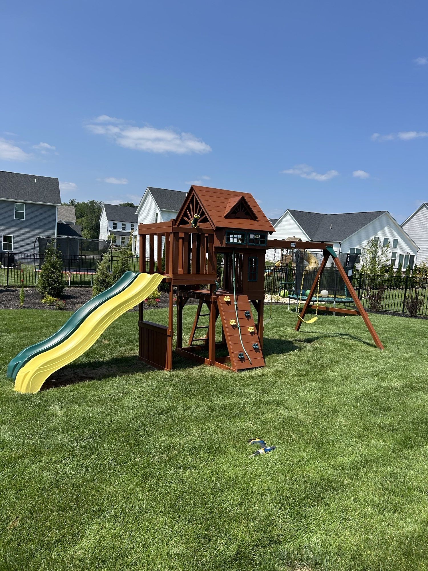 A wooden playset with a slide, climbing wall, and swing set on a green lawn in a suburban backyard.