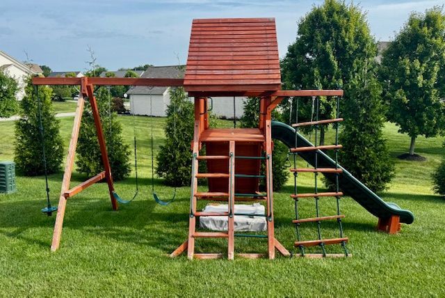 Wooden backyard playset with swings, ladder, climbing rope, and slide on green grass.