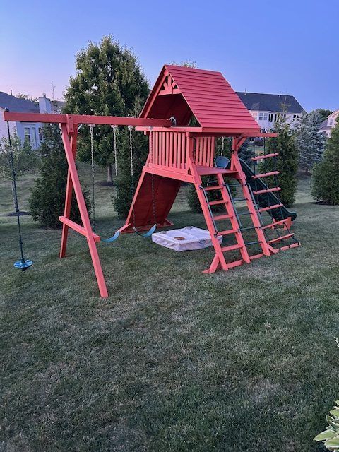 Red wooden playset with swings, slide, climbing wall, and sandbox on a grassy lawn.