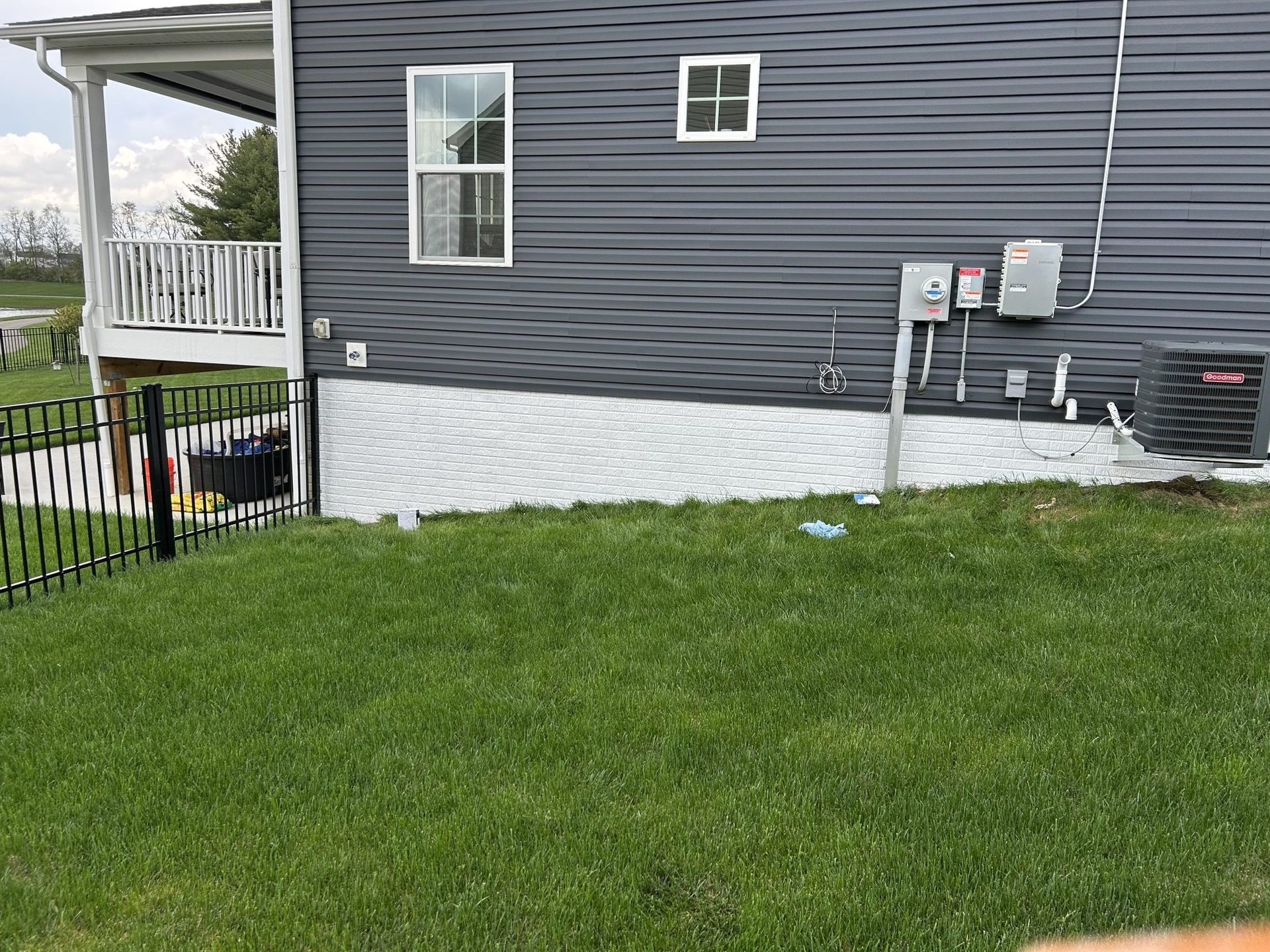 Side of a gray house with white patterned foundation, green grass, and black fence.